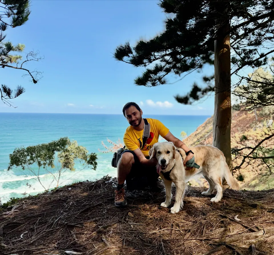Luka crouching beside Kala under the pines, with the sea behind them.